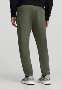 Oliwkowe spodnie o prostym kroju, wykonane z miękkiej tkaniny, z dwoma tylnymi kieszeniami, zestawione z szaro-czarnymi sneakersami.