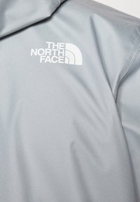 Harmaa takki, joka on valmistettu sileästä ja kevyestä kankaasta, jossa on valkoinen logo rinnassa. Mallissa on vetoketjullinen etuosa ja huppu.