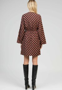 Femme portant une robe marron à rayures noires diagonales et à manches longues, associée à des bottes à talons noires remontant jusqu'aux genoux, se tenant de dos.