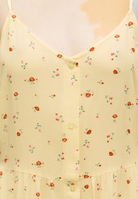 Κίτρινο φόρεμα με λεπτές τιράντες, μικρό κόκκινο και ροζ ανθισμένο σχέδιο και κουμπιά μπροστά κατά μήκος του κέντρου.