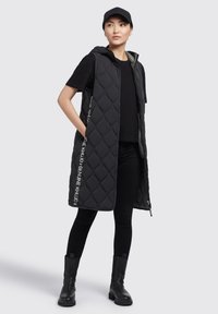 Schwarw gesteppte lange Weste mit Kapuze, seitlichen Taschen und weißem Text auf der Vorderseite. Trägt man über einem schwarzen Oberteil und schwarzen Leggings.