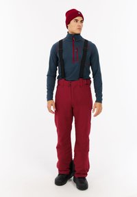 Rode sneeuwbroek met verstelbare schouderbanden, gecombineerd met een blauw langarmshirt met een ritskraag en zijaccentstiksels. Zwarte laarzen.
