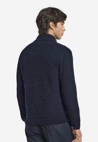 Pull en maille côtelée bleu marine avec un col montant et des manches longues, présentant un tissu texturé et un ourlet ajusté. Vue de dos.
