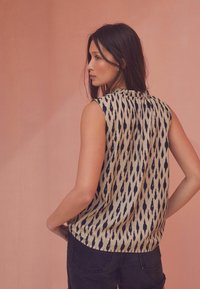 Ærmeløs bluse i beige med et sort ikat-mønster, der har plisseret ryg og rund kant, parret med mørke jeans.