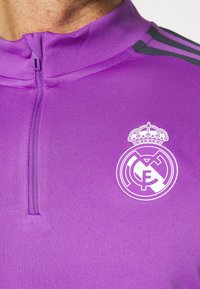 Lila sportig topp med kvartszip och hög krage, med svarta detaljer och en vit Real Madrid-logga på bröstet. Slät textur.