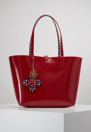 Rode glanzende totebag met lange hengsels en een blauw-rode bloemen sleutelhanger, tentoongesteld op een witte voetstuk.