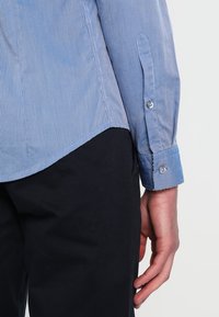 Modrá a bílá pruhovaná košile na knoflíky s dlouhými rukávy, bavlněný materiál a zaoblený spodní lem. Má manžety na knoflíky a hladkou texturu.