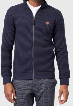 Homme portant une veste zippée texturée bleu marine sur une chemise blanche et un pantalon à carreaux gris, debout avec les bras détendus le long du corps.