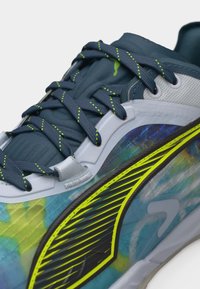 Κοντινό πλάνο ενός αθλητικού παπουτσιού σε μπλε και κίτρινο χρώμα, με σκουρό μπλε κορδόνια, αντανακλαστικές κίτρινες ρίγες και λεπτομέρειες από δίχτυ στο πάνω μέρος.