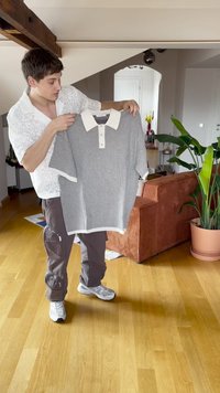 Pelēkā un baltā svītrota īsām piedurknēm polo krekls ar balto apkakli, ar pogu slēdzi un ribotām piedurknēm, ko tur cilvēks iekštelpās.