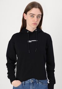 Hanorac negru cu un design ajustat, cu un logo alb pe piept, srtcuri pentru ajustare și mansete rigide. Asortat cu blugi albaștri cu talie înaltă.