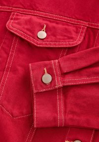 Red denim jacket fabric with contrasting yellow stitching, featuring a chest pocket and button closure detail. Smooth texture.
