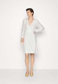 Vestido branco envolvente com tecido texturizado, mangas longas e decote em V profundo. Combinado com saltos em palha bege e uma pequena bolsa de ombro de tricô.