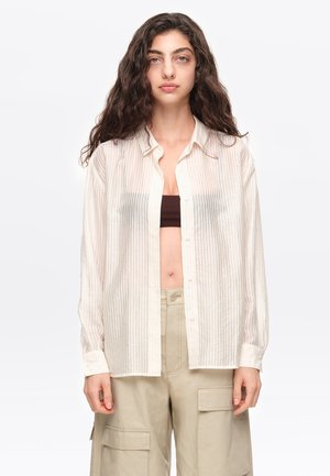 Mujer con cabello largo y rizado que lleva una camisa blanca transparente abotonada sobre un top corto negro y pantalones cargo beige, de pie frente a un fondo liso.