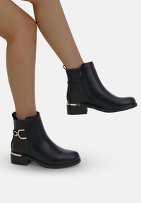 Bottines noires avec boucle dorée et accent sur le talon, portées sur des jambes devant un fond blanc uni, montrant des vues de côté et de dos.