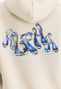 Crème hoodie met een grote, glanzende blauw-grijze grafische tekst "ALPHA" en "STUDIO EDITION" in lichtblauw, met een zachte, textuurstof.
