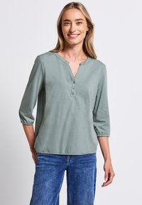 Blusa verde chiaro a maniche lunghe con scollatura a V e orlo elasticizzato; abbinata a jeans blu. Tessuto morbido, design casual.