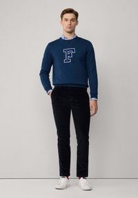 Felpa blu marina con design della lettera bianca "F", abbinata a pantaloni in corduroy neri e sneakers bianche. Sopra, indossa una camicia colletto azzurra chiara.