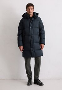 Granatowa kurtka puchowa z kapturem, z pikowanym wzorem, zatrzaskami i bocznymi kieszeniami. Noszona na ciemnoszarych spodniach i czarnych butach.