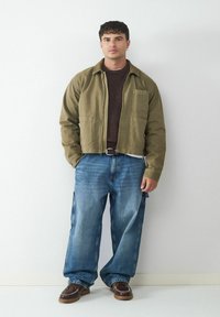 Olive green cropped jacket over a dark brown sweater, paired with light blue baggy jeans and brown shoes with white stitching details.