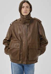 Hnedá kožená bunda s vysokým golierom, veľkými prednými vreckami a ramennými epolety. Hladká textúra a voľný strih dopĺňajú dizajn.
