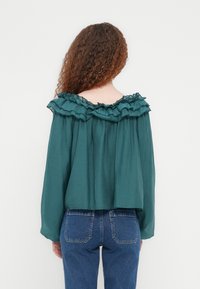 Πράσινο μπλουζάκι με άνοιγμα στους ώμους και βολάν στον λαιμό, φαρδιά γραμμή, μακριά μανίκια και μαλακή υφή, σε συνδυασμό με ψηλόμεσο τζιν μπλε χρώματος.