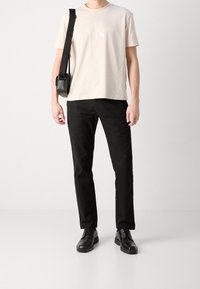 T-shirt in cotone beige chiaro con colletto tondo e logo sottile, abbinata a pantaloni dritti neri e scarpe in pelle nere.