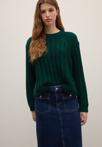 Young woman with long wavy hair wearing a dark green knit sweater and high-waisted dark blue denim skirt against plain background.