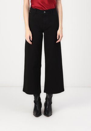 Zwarte wijde broek van katoen, met een knoopsluiting, frontzakken en een gladde textuur. Gedragen met zwarte haklaarzen.