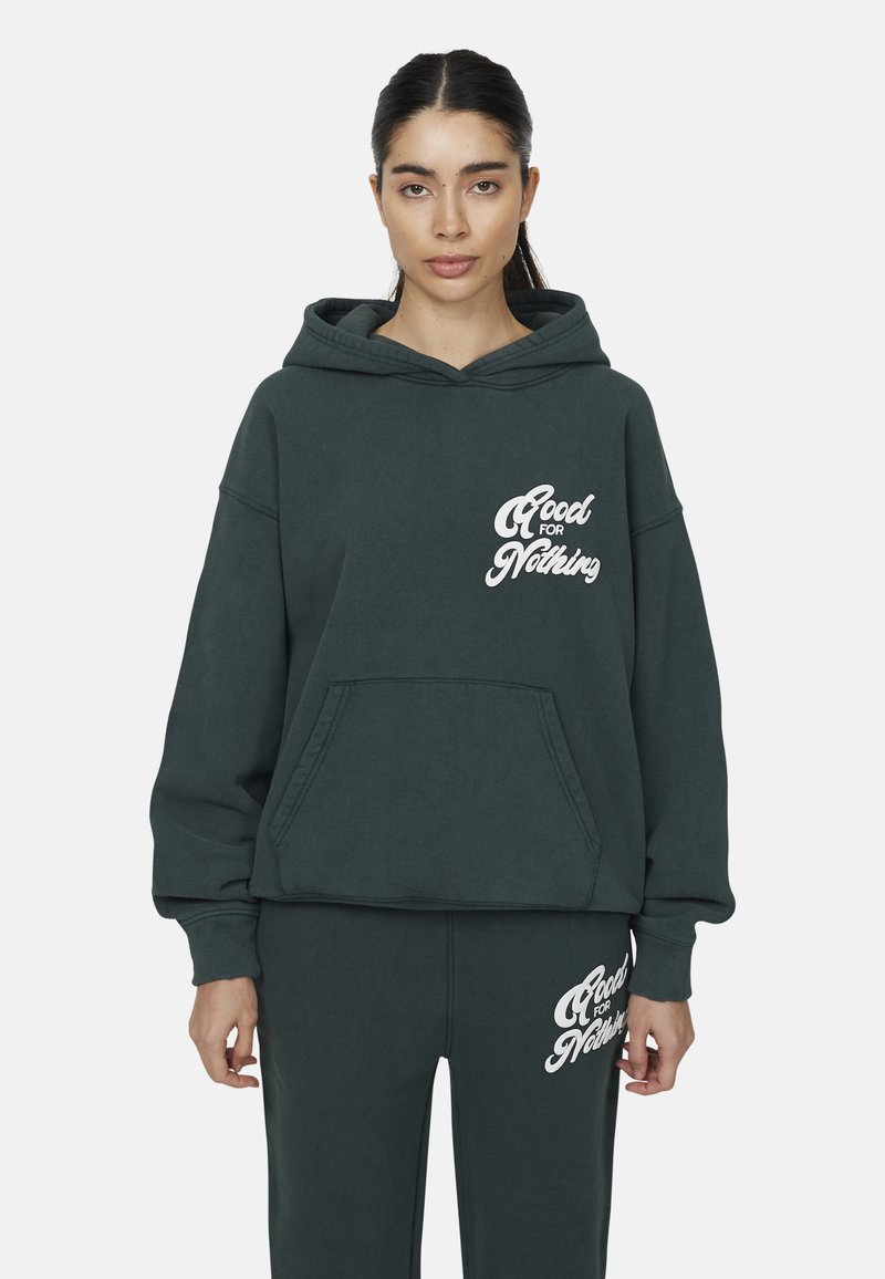 Felpa verde scuro con tasca frontale, con la scritta bianca "Good for Nothing." Abbinata a pantaloni coordinati, che mostrano anch'essi la stessa scritta.