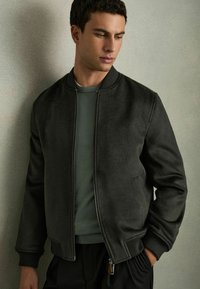 Tumes roheline bomber-jakk, millel on lukk ees, elastsed mansetid ja küljetaskud. Stiliseeritud sobiva rohelise ümarakaeluse särgi peal. Pehme tekstuur.