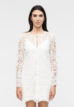 Γυναίκα που φοράει ένα λευκό δαντελωτό μακρυμάνικο φόρεμα με λαιμόκοψη τύπου κλειδαρότρυπα και περίπλοκα λουλουδένια σχέδια, στέκεται μπροστά σε απλό φόντο.