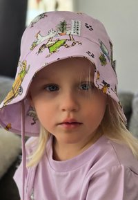Rosa bucket hat med lekfulla djur- och tecknad serie-mönster; bomullsmaterial; bred brätte. Matchas med en ljus lila t-shirt.