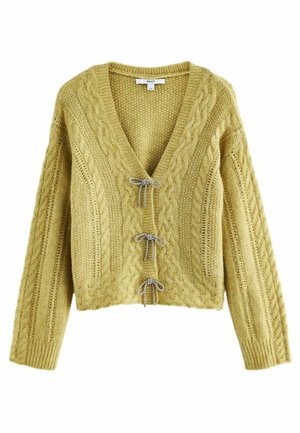 Personne portant un cardigan court jaune en maille torsadée avec trois nœuds noués, un jean large gris foncé, et des chaussures argentées à bouts pointus.