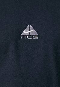 Dunkelblaues T-Shirt mit einem gestickten Nike ACG-Logo in Silber. Hergestellt aus einem glatten, strukturierten Stoff mit einer figurschmeichelnden Passform.