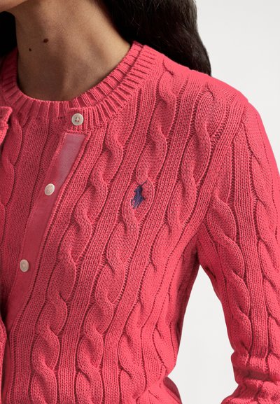 Spilgti rozā krāsas džemperis ar pinumu rakstu, ar ribota auduma apkakli. Aprīkots ar pogas aizdares plāksni un nelielu tumši zilu logotipu. Mīksts tekstūra, viegls dizains.