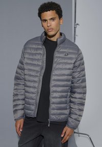 Noor mees, kes kannab musta särgi üle halli vatitekki jope, seisab tavalise tausta ees lõdvestunud asendis.