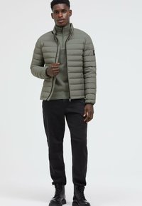 Puffer jacket in olive green with horizontal quilted design, black accents, high collar, paired with dark trousers and black boots.
