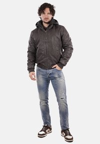 Giubbotto bomber marrone scuro con cappuccio, chiusura lampo frontale, polsini a coste, indossato con jeans chiari strappati e sneakers marroni.