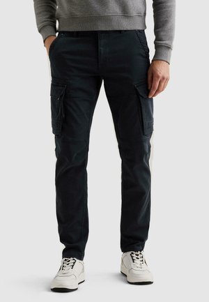 Mann trägt schwarze Cargohosen mit mehreren Taschen und weiße Sneakers, steht vor einem schlichten Hintergrund.