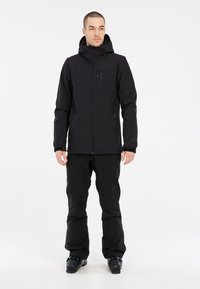 Veste de ski noire isolée avec capuche, poche poitrine zippée et poches latérales ; associée à un pantalon noir assorti et des bottes de ski.