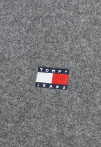 Grauer Stoff mit einer weichen Textur weist einen rechteckigen gestickten Patch auf. Der Patch hat navyblaue, weiße und rote Abschnitte mit dem Text "TOMMY JEANS".