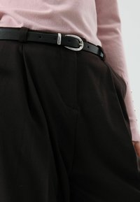 Ceinture noire avec une boucle argentée, portée sur un pantalon foncé. Le haut est un pull à manches longues rose clair avec des boutons en détail sur les manches.