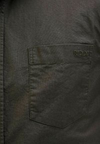 Tessuto della camicia verde scuro con una texture liscia, dotato di una sola tasca sul petto e logo ricamato "R.D.D." nello stesso colore.