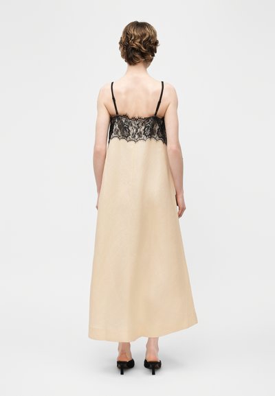 Γυναίκα φαίνεται από πίσω, φορώντας ένα μπεζ φόρεμα μέχρι τον αστράγαλο με μαύρη δαντέλα στο πάνω μέρος και λεπτές τιράντες, συνδυασμένο με μαύρα παπούτσια με τακούνι.