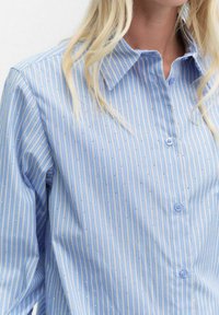 Jasnoniebieska koszula z guzikami, w pionowe paski z małym białym wzorem. Materiał jest gładki o klasycznym kroju kołnierza.