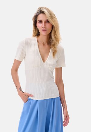 Weißes geripptes Strickoberteil mit tiefem V-Ausschnitt und kurzen Ärmeln, kombiniert mit lockeren hellblauen Hosen. Strukturiertes Gewebe und klares Design.