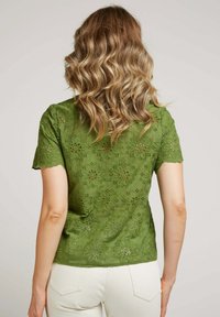 Haut en dentelle verte avec broderie florale et ourlet festonné, à manches courtes et coupe ajustée. Le dos est principalement opaque.