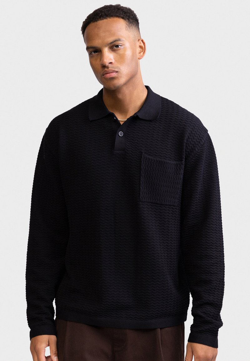 Pulôver preto com padrão texturizado ondulado e um pequeno bolso no peito. Apresenta gola com colarinho e mangas compridas.