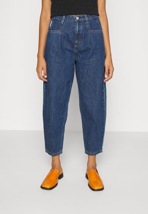 Persona que lleva vaqueros azules de talle alto y corte holgado, recortados por encima de los tobillos, y zapatos deportivos naranja brillante sin cordones con suelas negras.